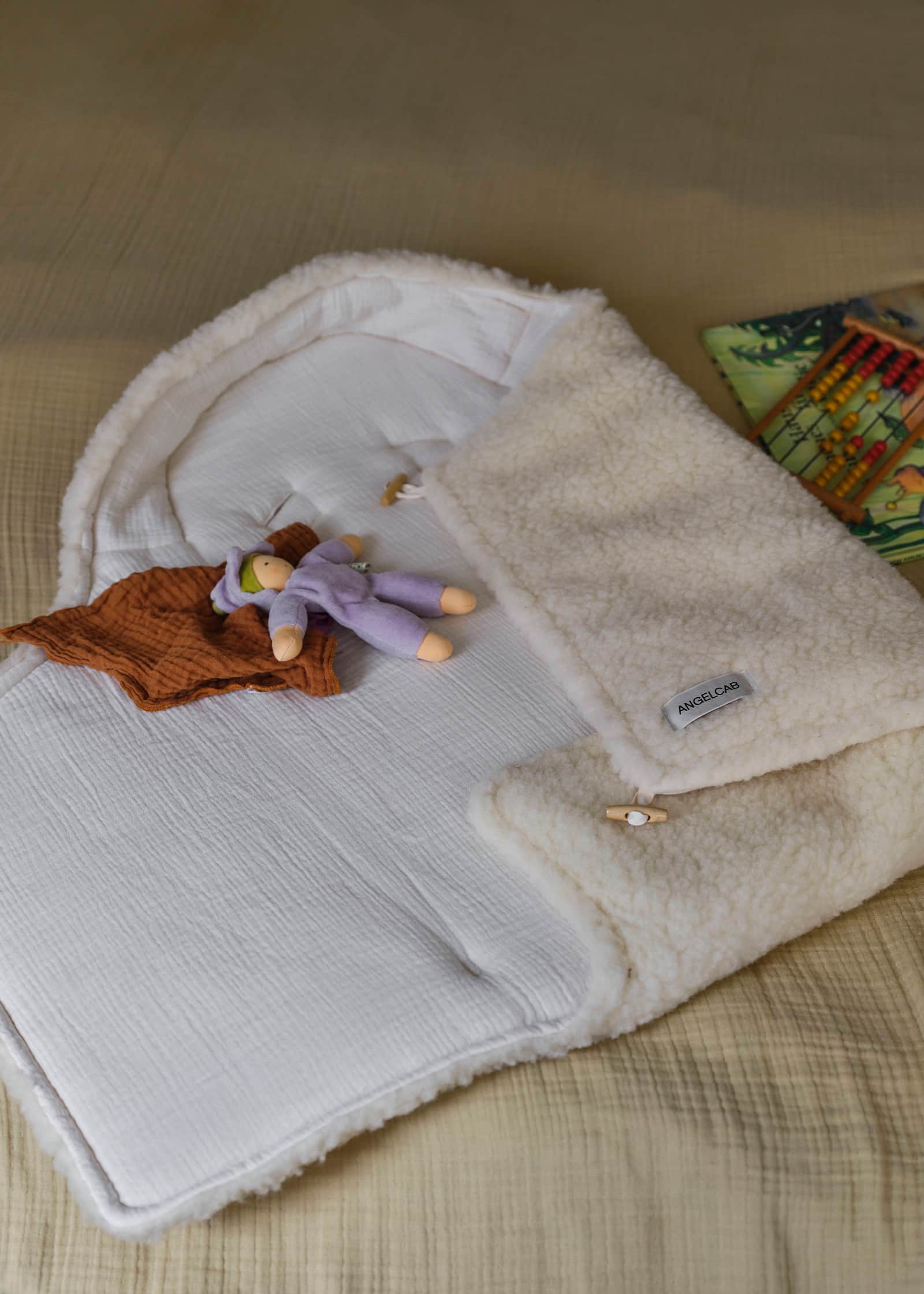 Geöffneter ANGELCAB Babyschlafsack aus Wollplüsch mit Musselin-Innenfutter, Holzknopf und Stoffpuppe auf einem Bett