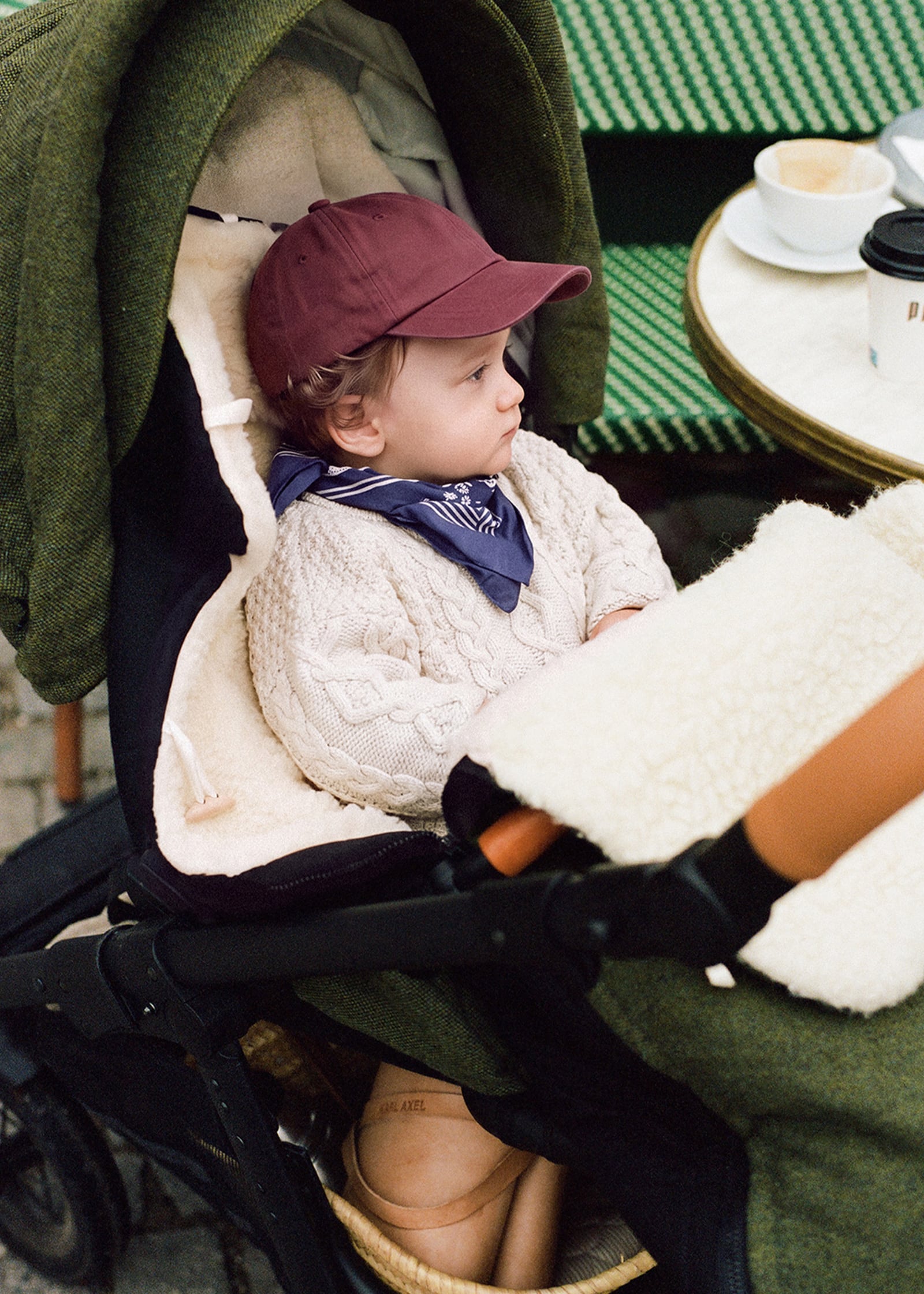 Kleinkind im Sportsitz eines ANGELCAB Kinderwagens mit grünem Tweed-Verdeck, Lammfell-Auflage und Fußsack im Café